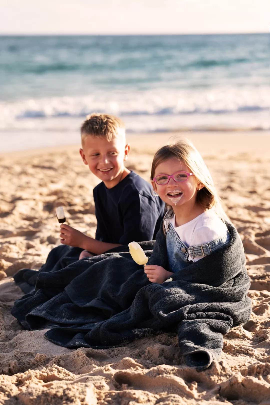 Two children wrapped in a beach towel eating icy poles on the sand, with the ocean behind them