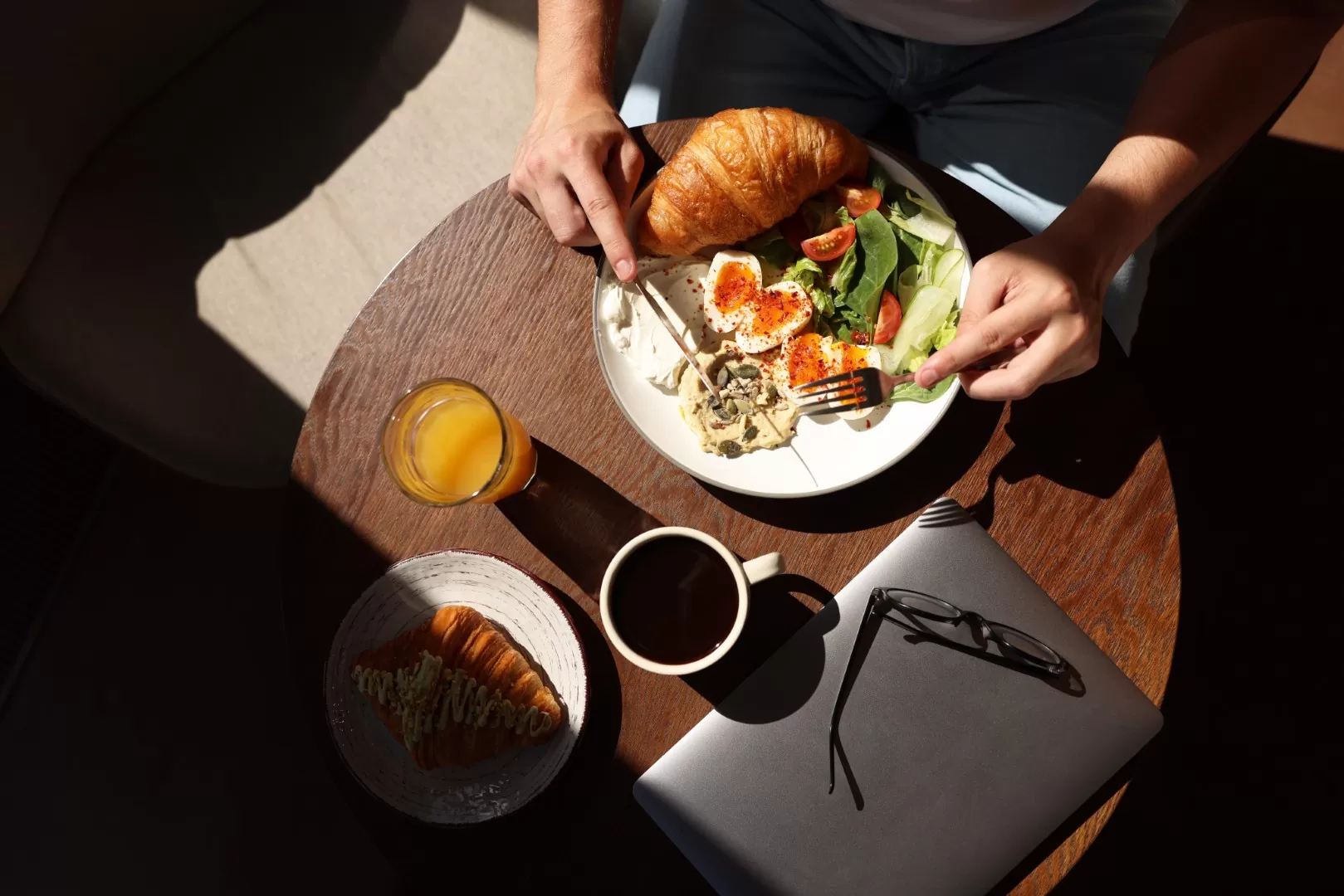 Overhead view of a breakfast table with a croissant, salad and coffee, with a laptop and glasses nearby
